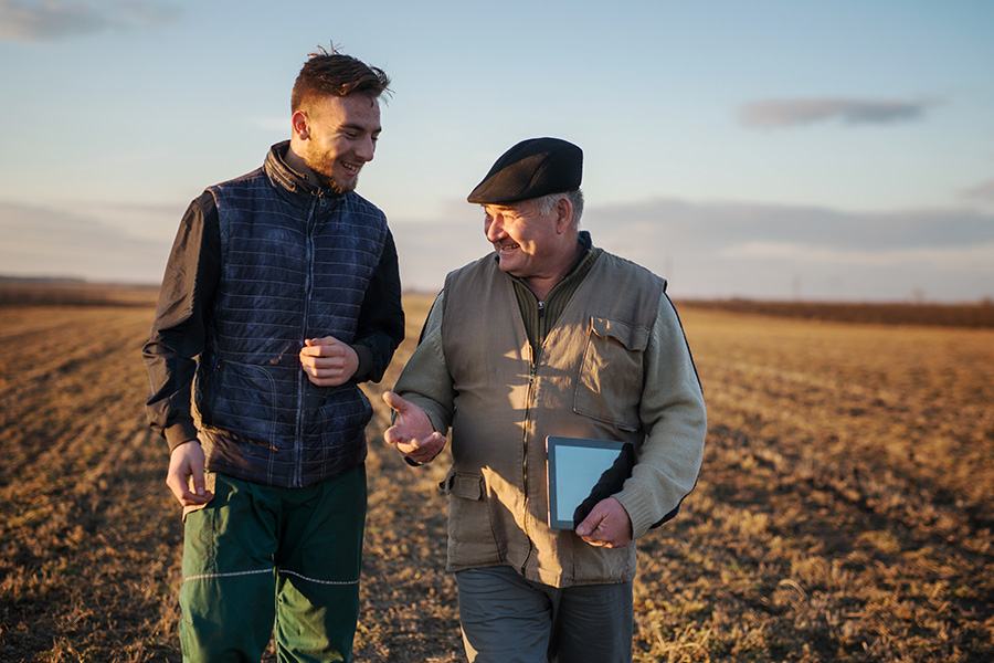 About Our Agency - Father and Son Farmers Standing Together on a Field Smiling and Talking With Each Other During Sunrise