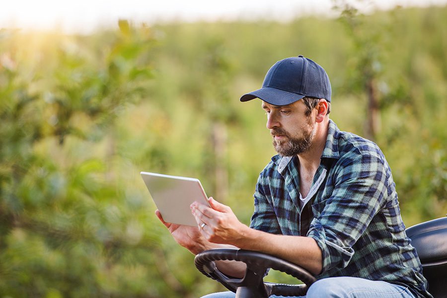 Client Center - A Farmer With His Tablet Sitting on a Mini Tractor Outdoors in His Orchard